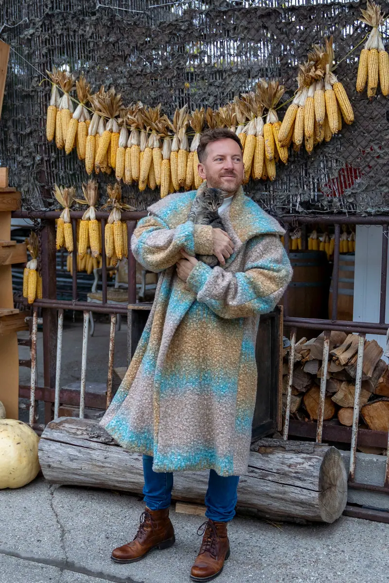 Lifestyle fotenie — muž v pestrom pletenom kabáte pred stenou so sušenou kukuricou, exteriér