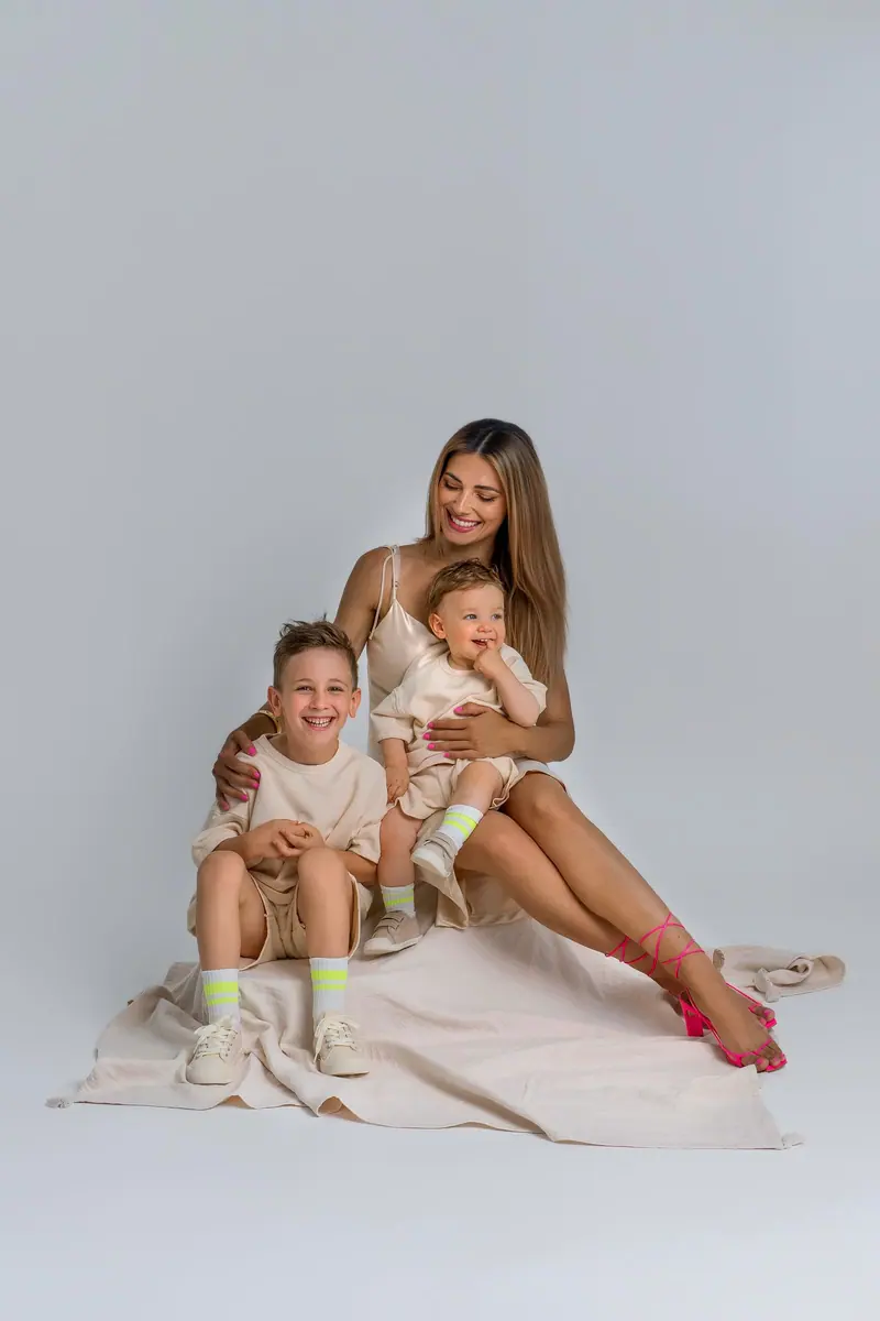 Fotenie mama a baby — mama sedí s dvomi synmi na deke, sivé pozadie, široký záber