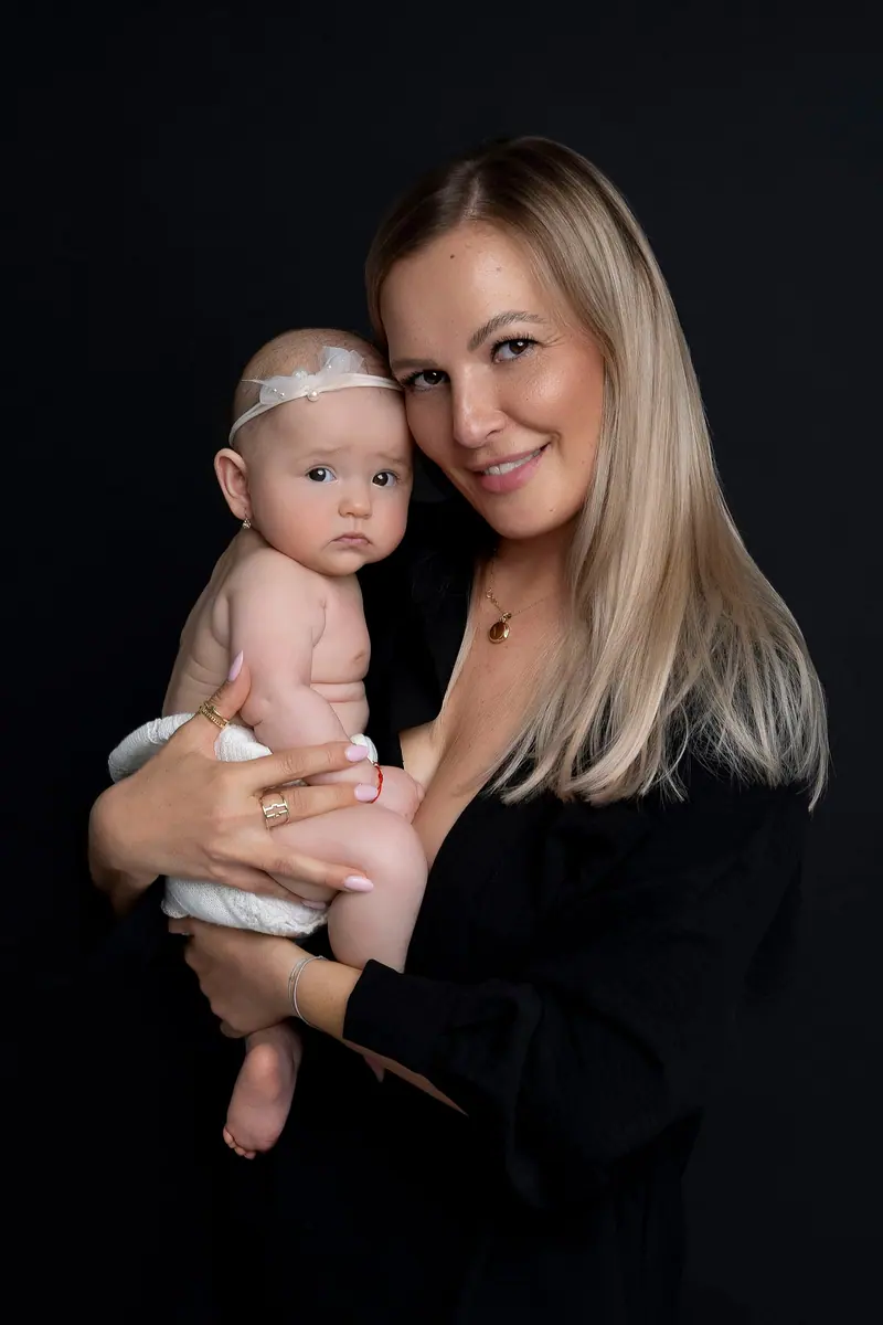 Fotenie mama a baby — blond mama drží bábätko s čelenkou, čierne pozadie