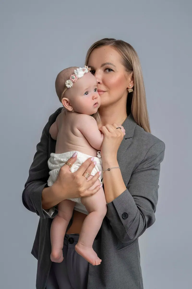 Fotenie mama a baby — blond mama v sivom saku drží bábätko, sivé pozadie