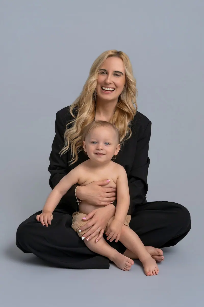 Fotenie mama a baby — blond mama v čiernom sedí s bábätkom v lone, sivé pozadie