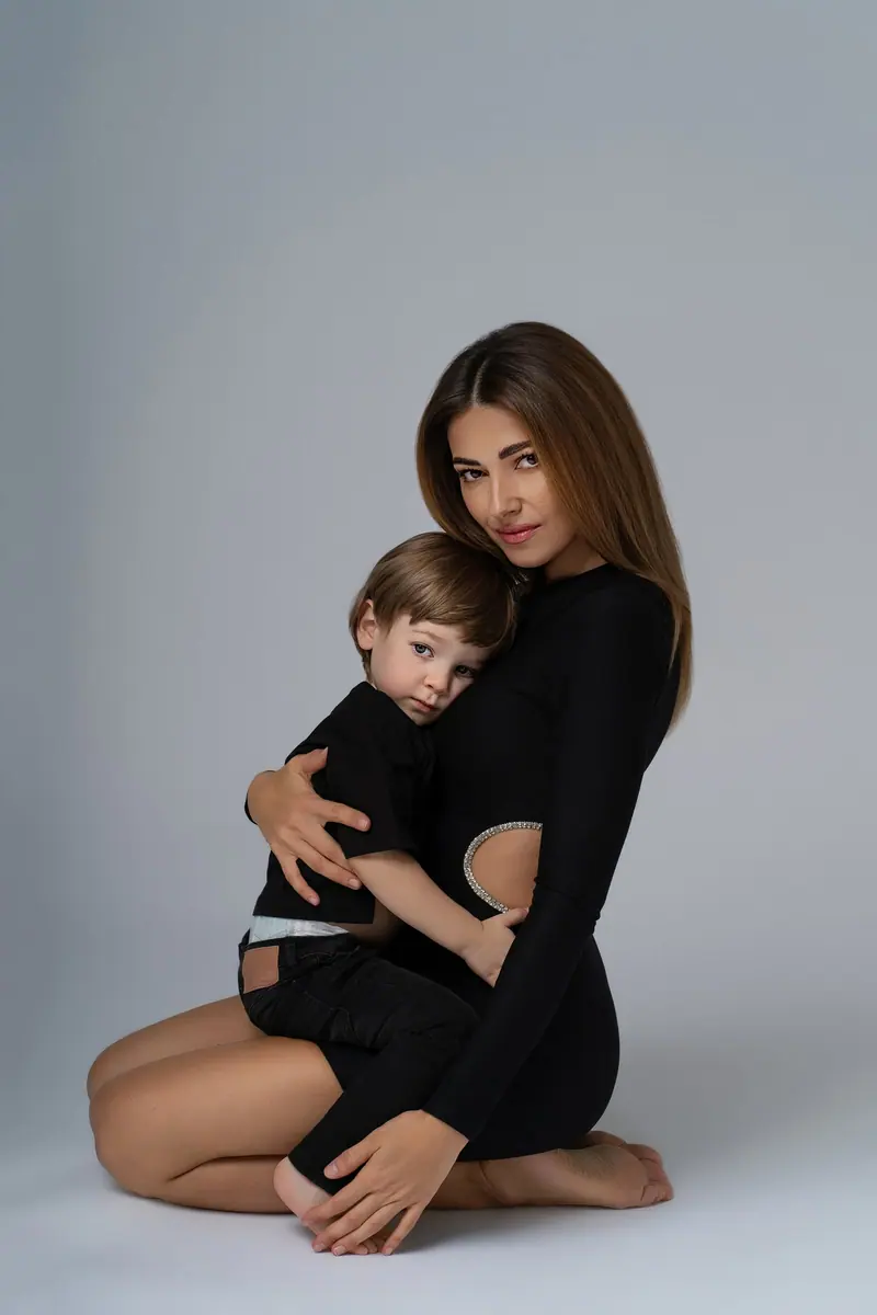 Fotenie mama a baby — mama v čiernom kľačí s dieťaťom v objatí, sivé pozadie