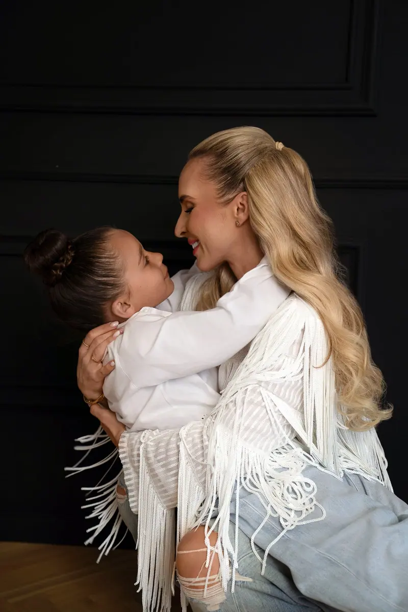 Fotenie mama a baby — blond mama v bielom so strapcami objíma dcérku, tmavé pozadie