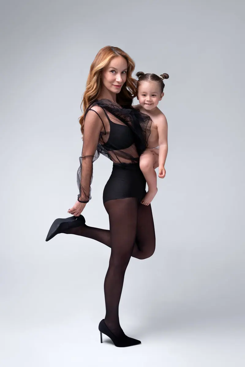 Fotenie mama a baby — mama v čiernom stojí na jednej nohe, dcérka s dvoma drdolčekmi na ruke