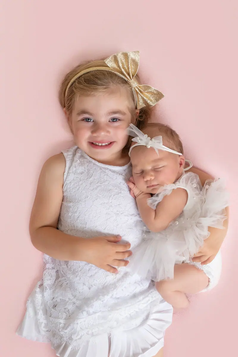 Newborn fotenie — sestra drží novorodenca, ružové pozadie, zhora