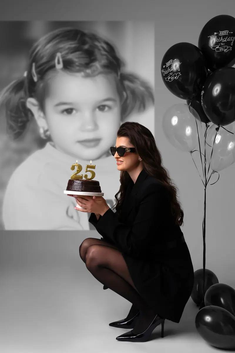 Narodeninové fotenie — žena v čiernom s tortou „25“, čierne balóny a portrét z detstva v pozadí