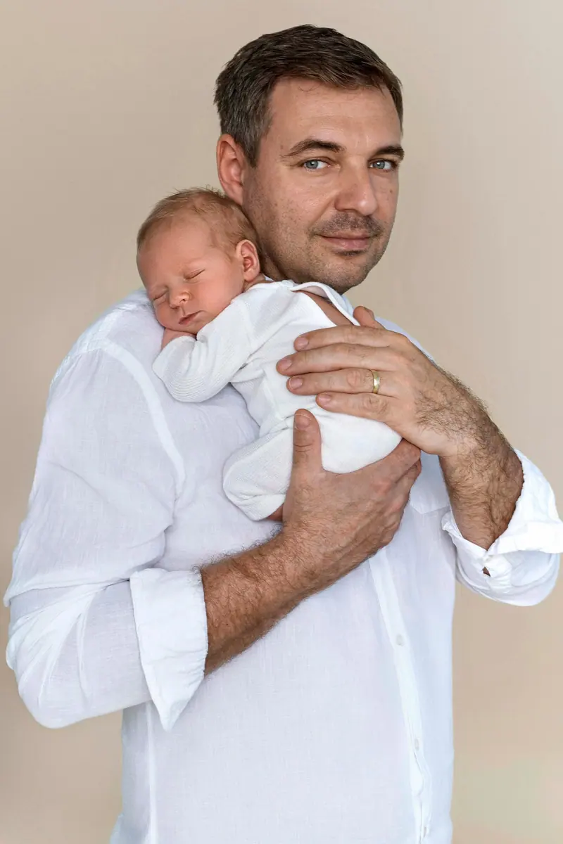 Newborn fotenie — otec v bielej košeli drží novorodenca, béžové pozadie