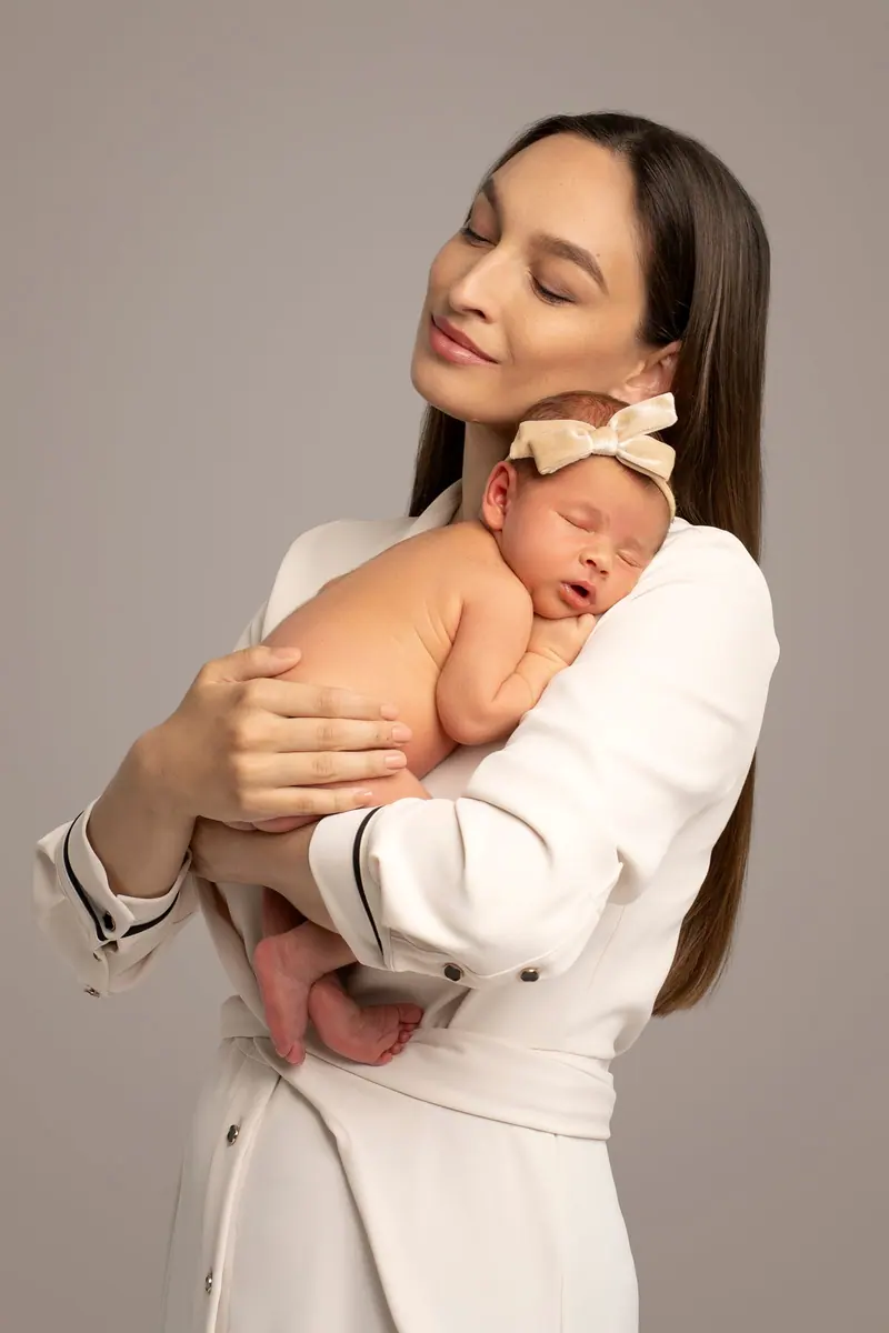 Newborn fotenie — mama v krémovom drží novorodenca, sivé pozadie