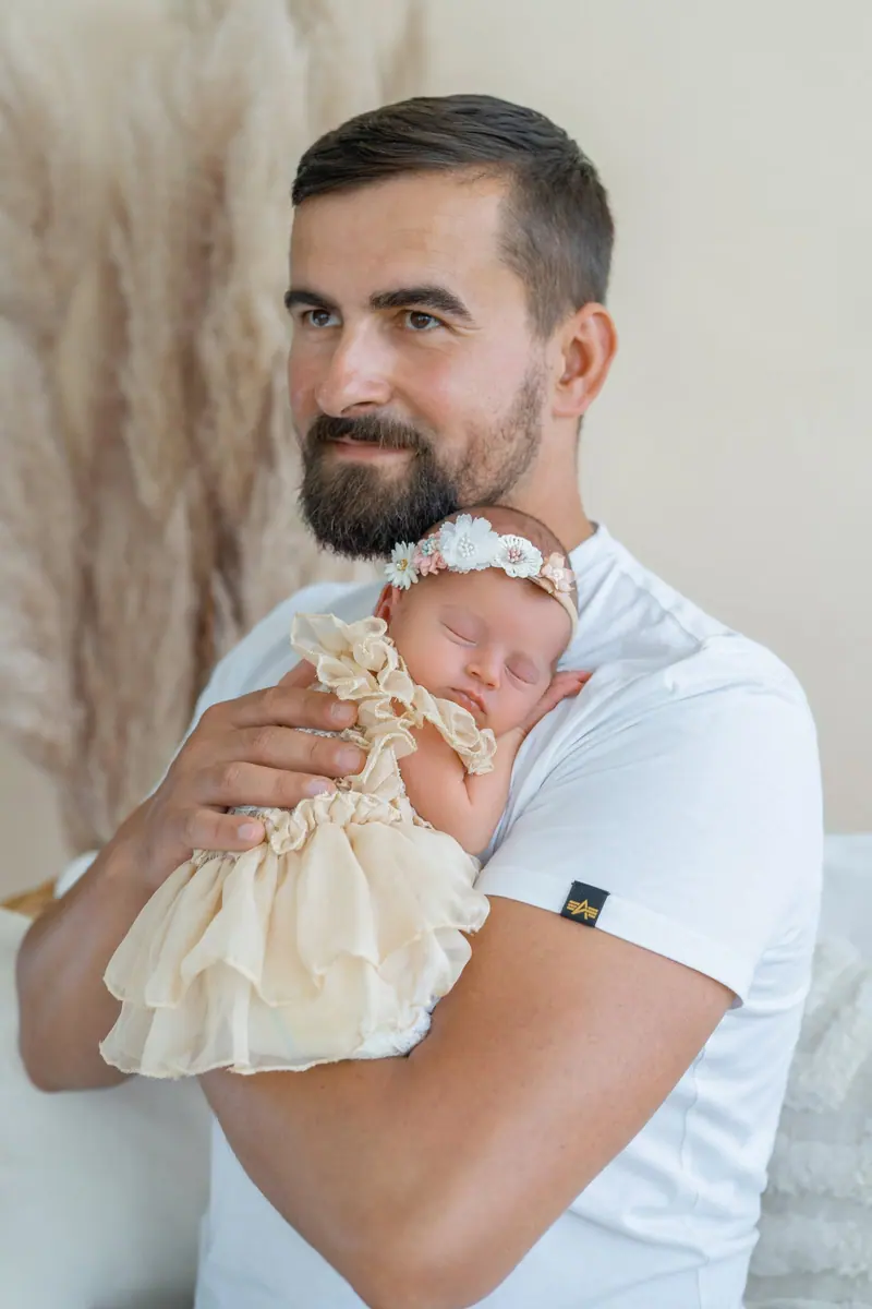 Newborn fotenie — otec v bielom tričku drží spiace bábätko v krémových šatách