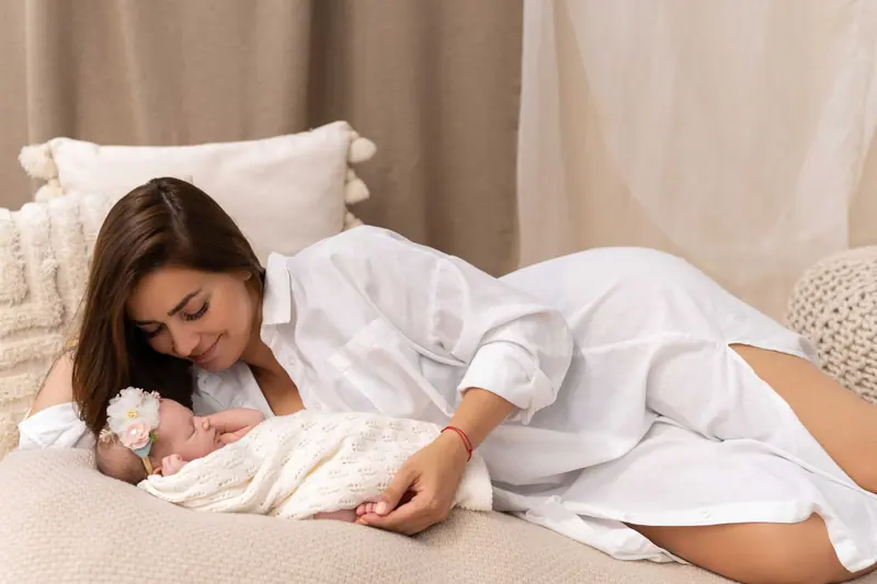 Newborn fotenie — mama v bielej košeli leží pri bábätku, béžové záclony