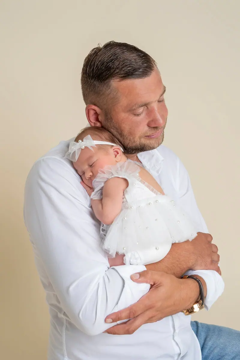 Newborn fotenie — otec v bielej košeli drží bábätko v bielych šatách