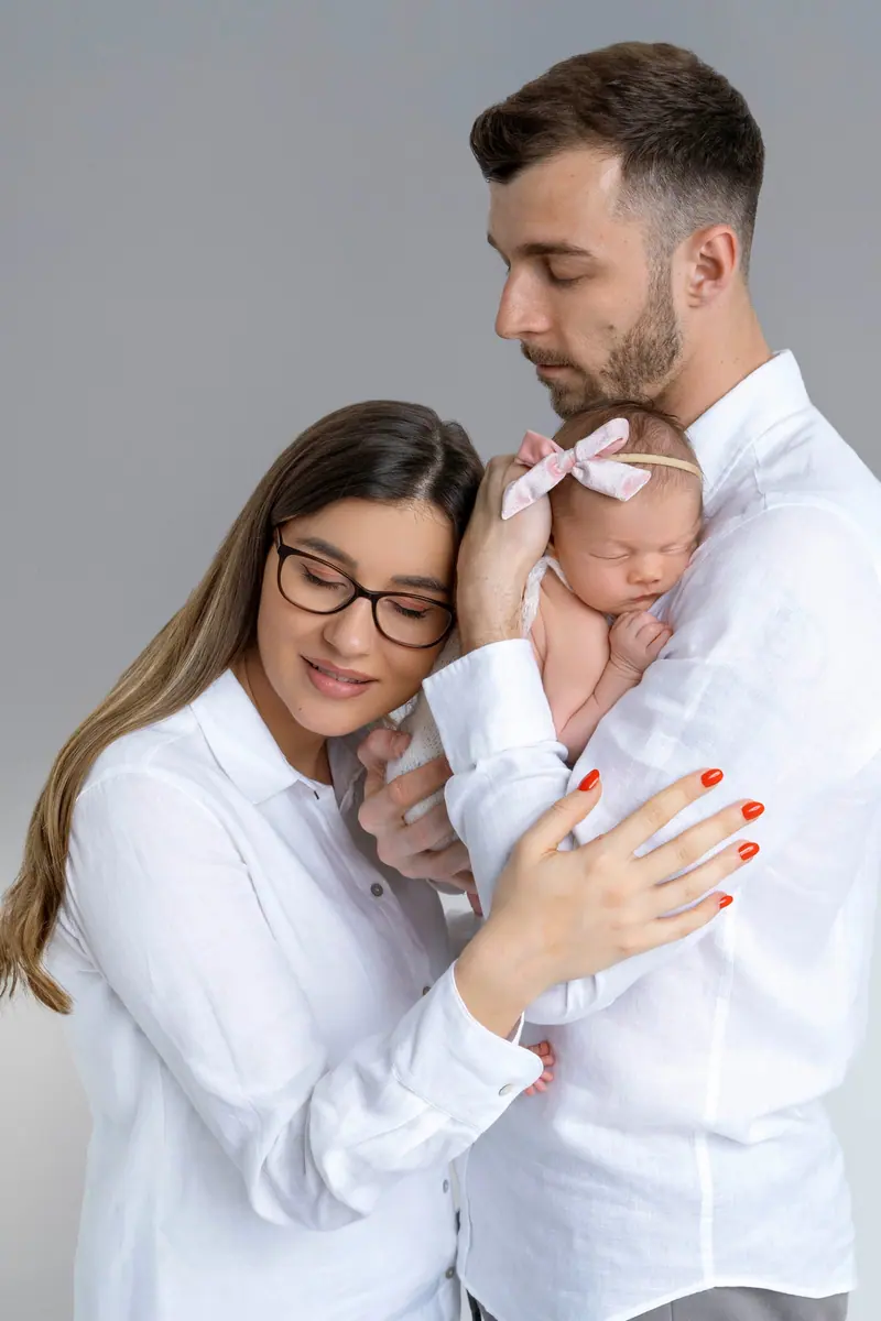 Newborn fotenie — rodičia v bielych košeliach s novorodencom s ružovou mašľou
