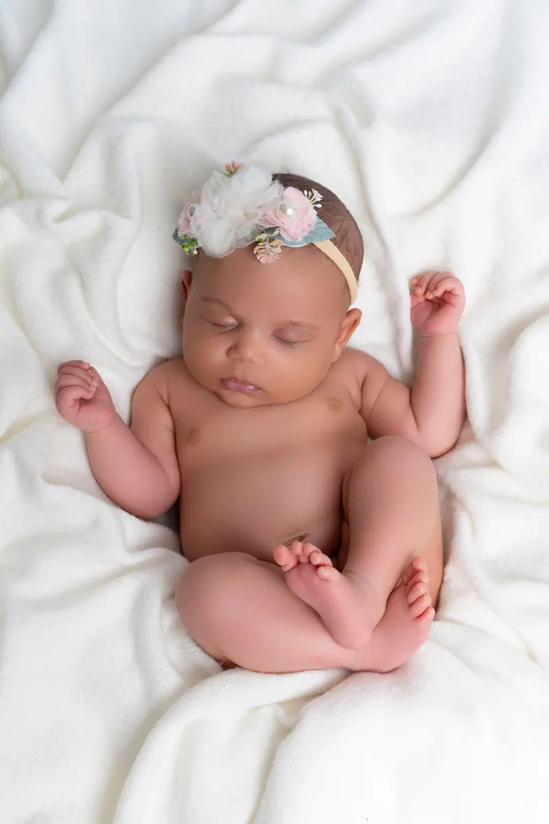 Newborn fotenie — novorodenec s kvetinovou čelenkou leží na bielej deke, záber zhora