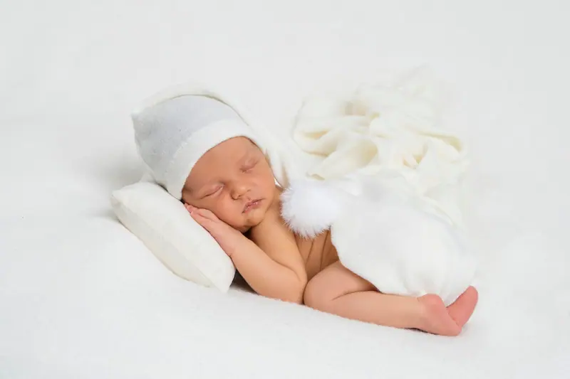 Newborn fotenie — novorodenec v bielej čiapke s brmbolcom na vankúšiku