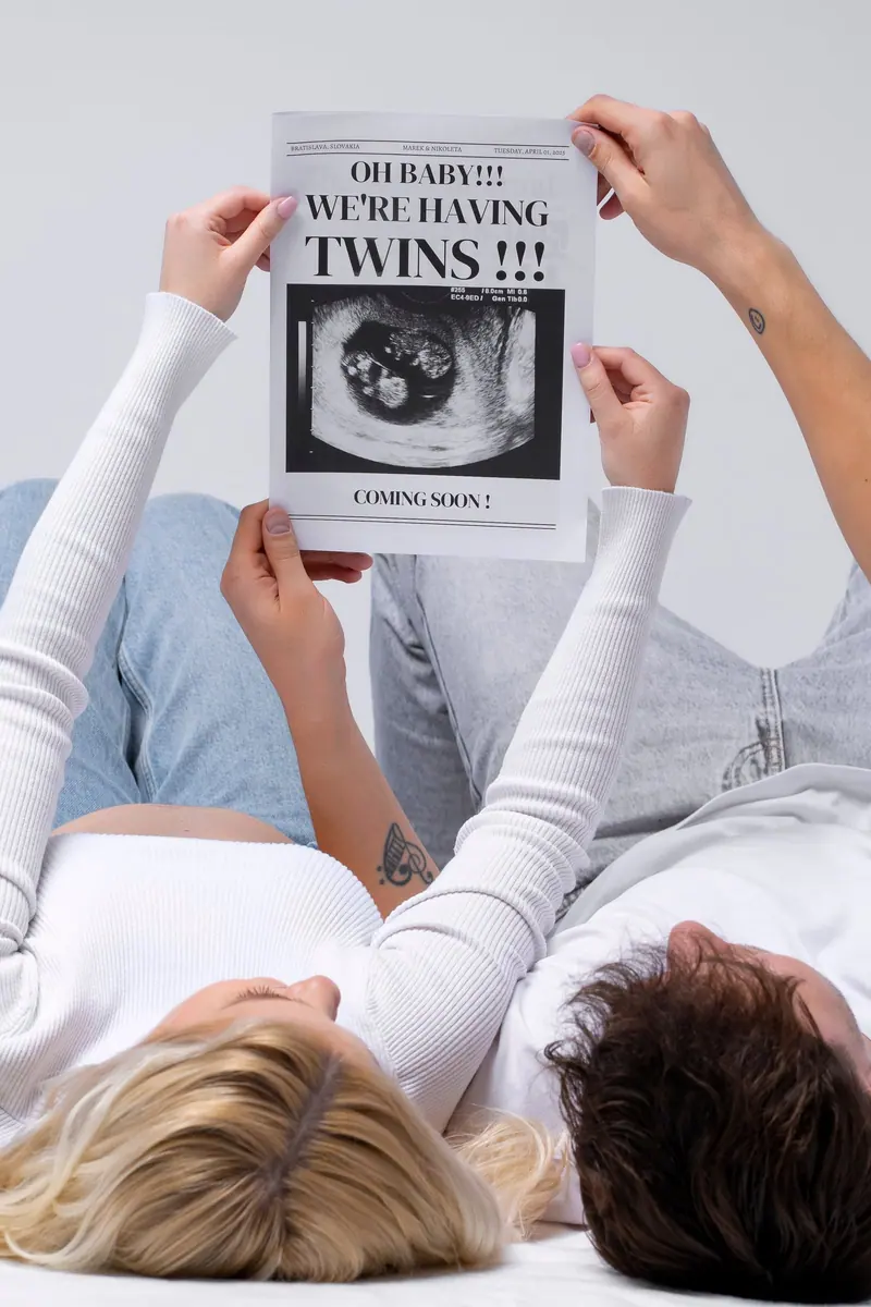 Tehotenské fotenie — ruky držia noviny „We’re having TWINS“, pár leží